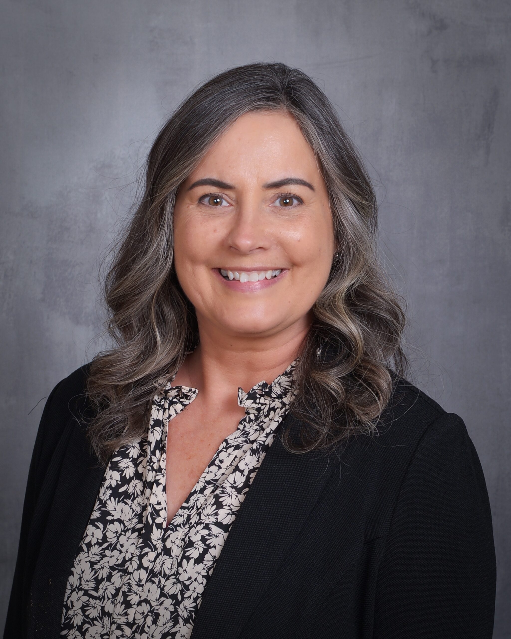 Smiling woman with wavy, gray hair wearing a black blazer over a floral blouse, against a gray backdrop. Suitable for a professional profile.