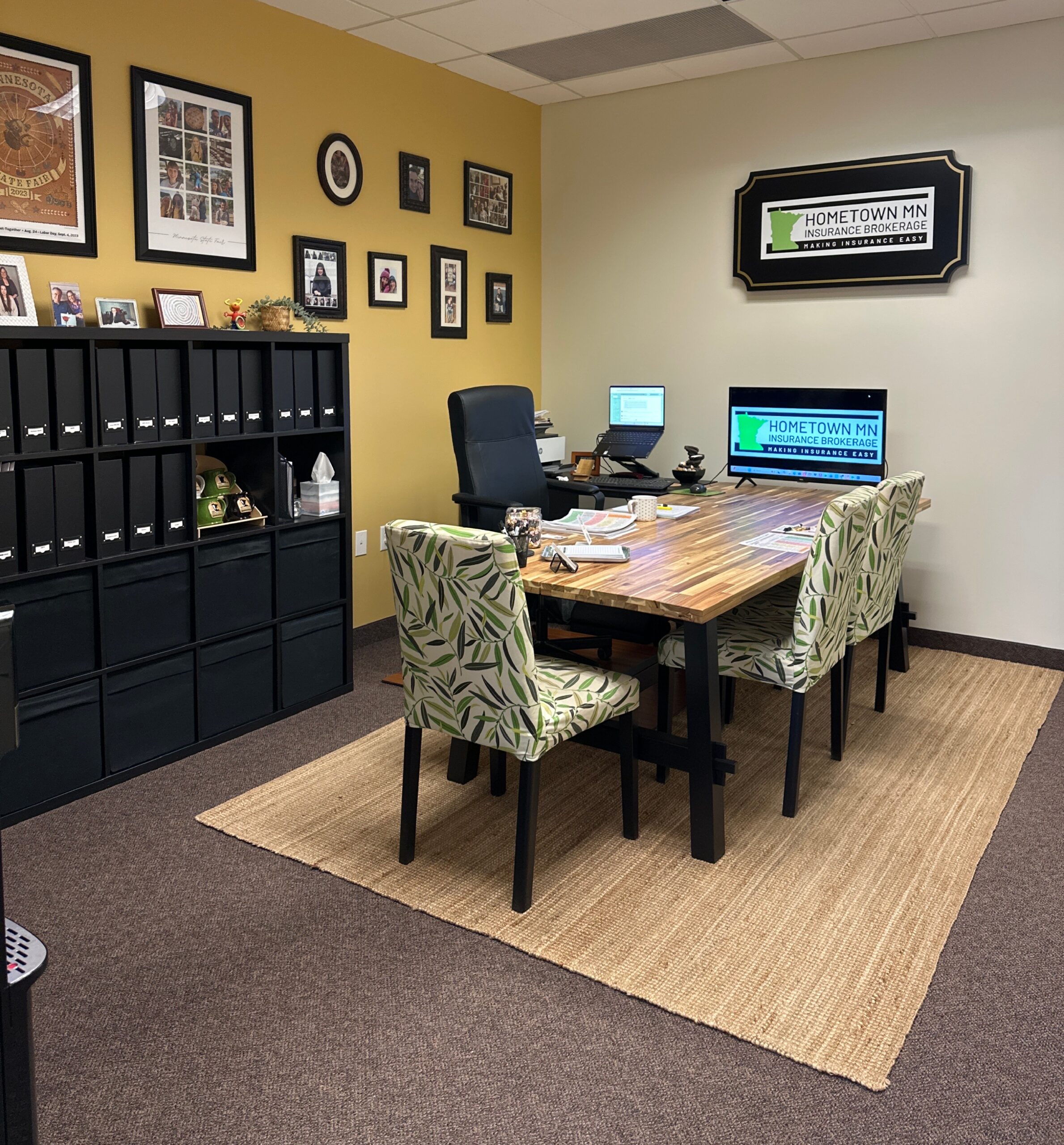 Modern office space featuring a wooden table surrounded by patterned chairs, a computer desk, and framed photos on the yellow wall.