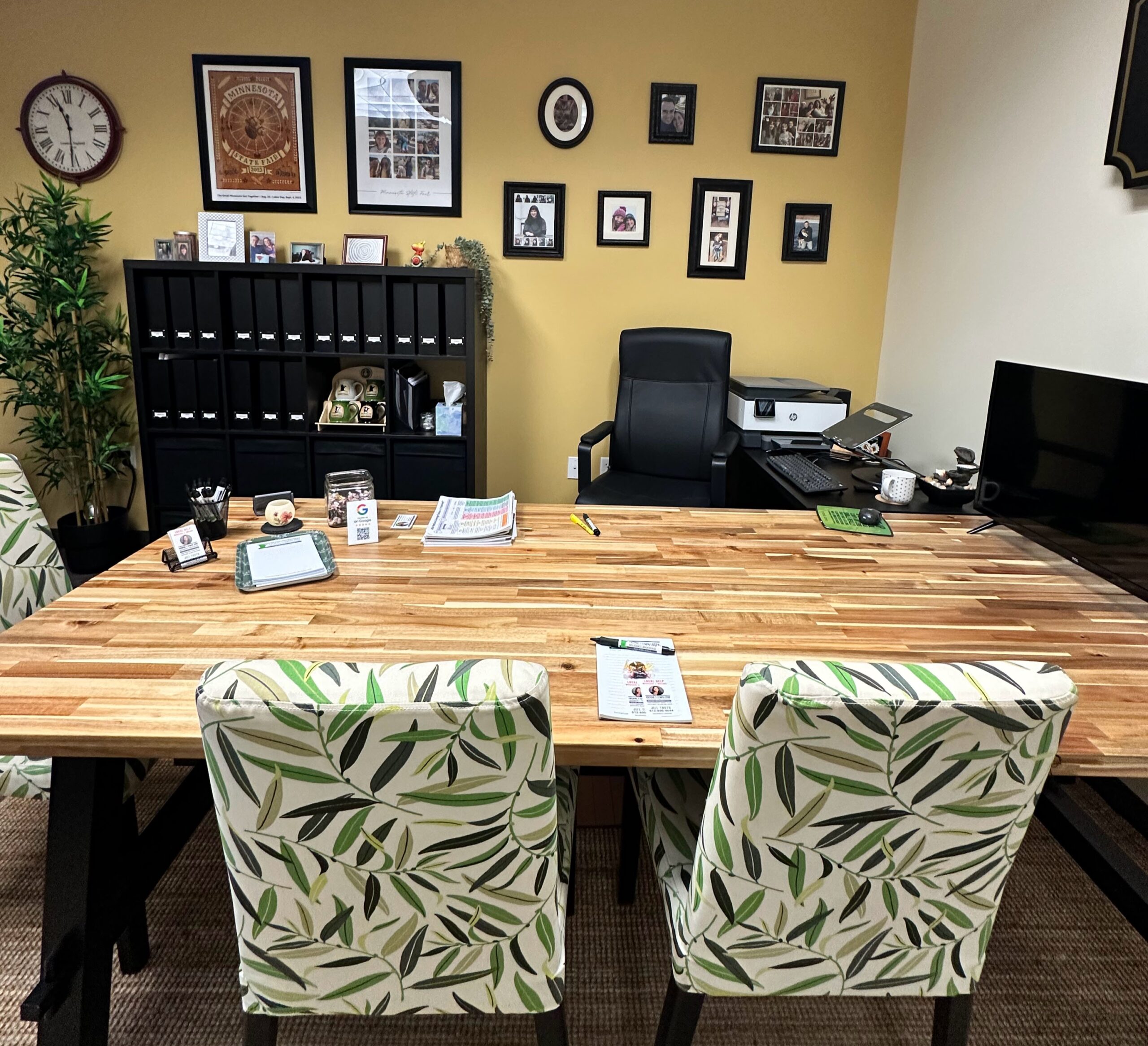 Bright office space with a wooden table, floral chairs, and a black executive chair. Organized workspace features decorative elements and documents.