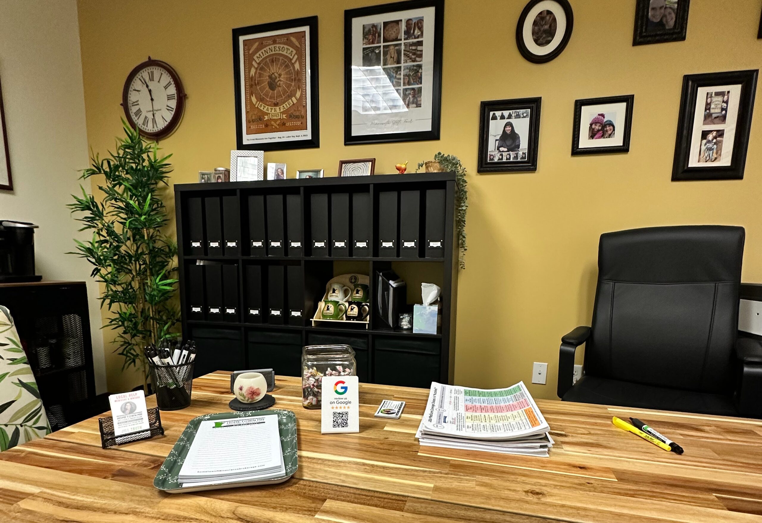 A well-organized office space featuring a wooden desk, filing cabinets, and framed photos. Essentials like a clock, documents, and office supplies are visible, reflecting a professional atmosphere.